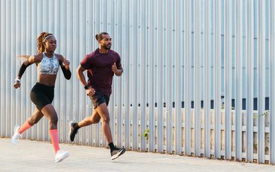 Volledig lichaam van zelfverzekerde gemotiveerde jonge Afro-Amerikaanse mannelijke en vrouwelijke hardlopers in sportkleding die samen trainen en snel rennen op de stoep bij het hek tijdens buitentraining