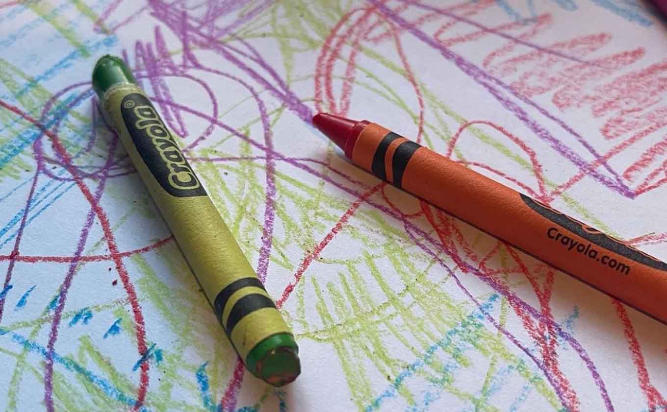 Een groen krijtje en oranje krijt zitten bovenop een wit stuk papier bedekt met kleurrijke krabbels.