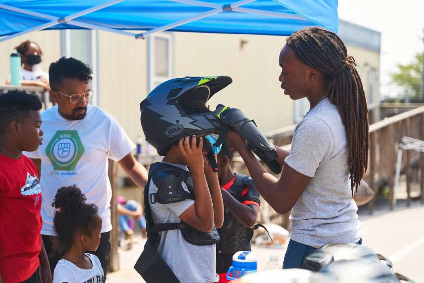 Een vrouw helpt een kind om een helm te dragen terwijl kinderen en een volwassene op de achtergrond spreken