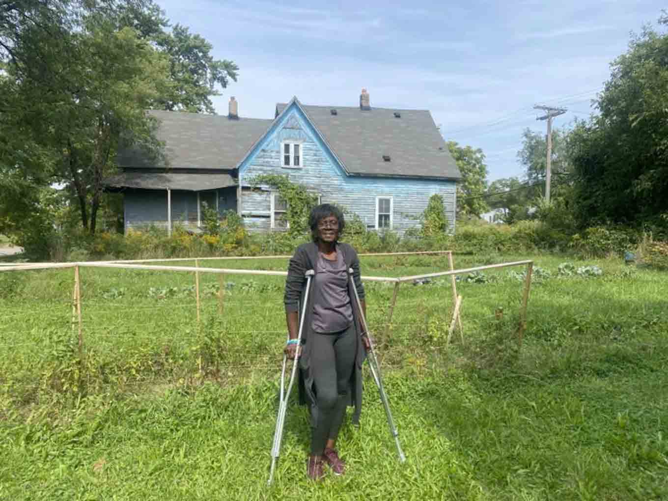 Pamela poseert voor de camera omringd door groen en een huis op de achtergrond.