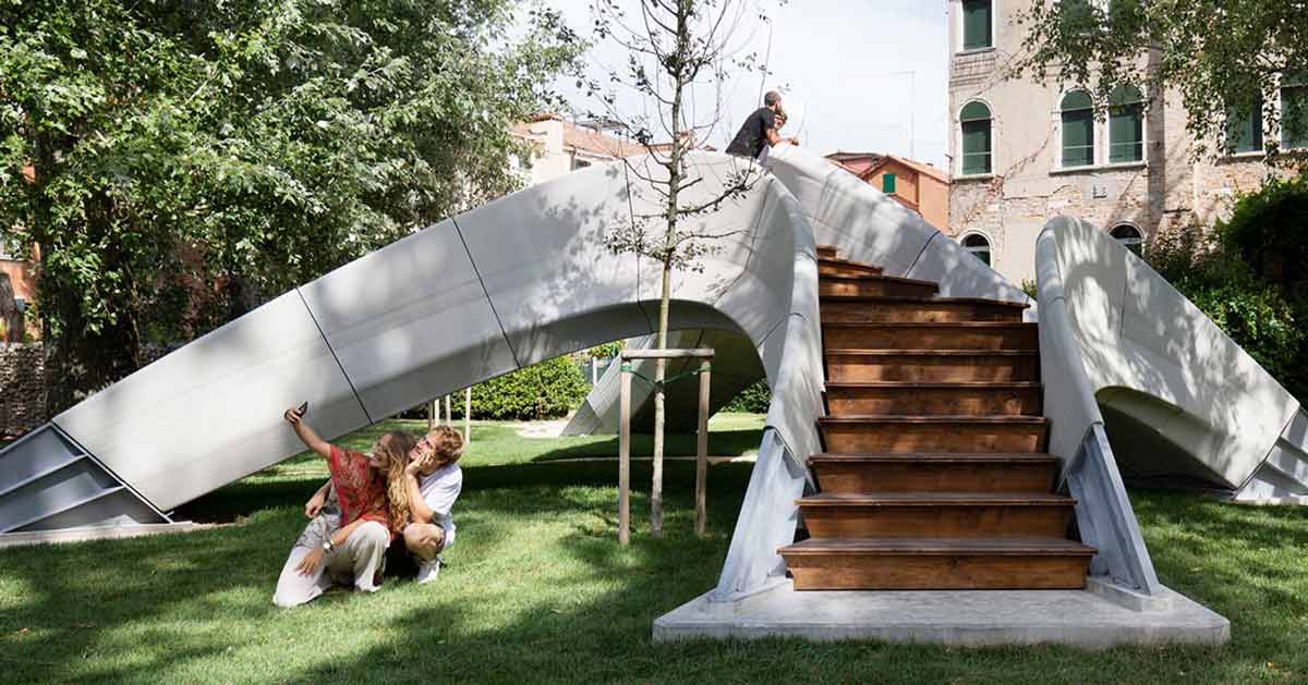 Een kleine unieke brug zit midden in een park. Het is beton en een stel maakt er een selfie voor. De trappen zijn gemaakt van hout.