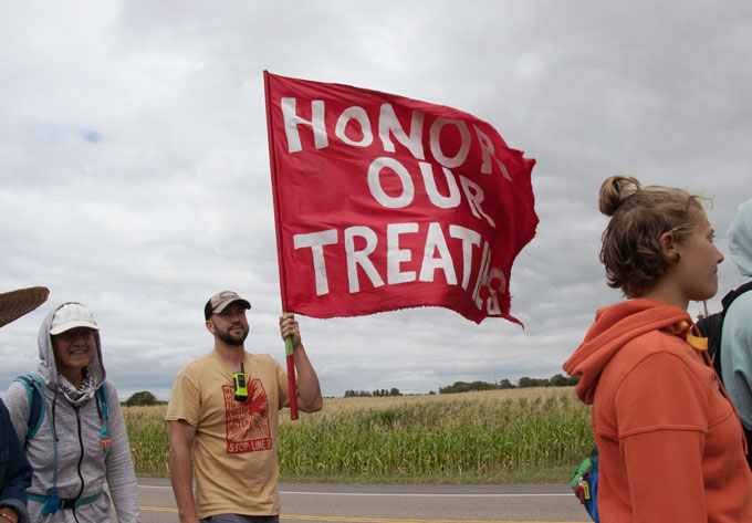 Een man marcheert met een menigte langs de kant van een landelijk gebied weg, met een rode vlag met de tekst EER ONZE VERDRAGEN