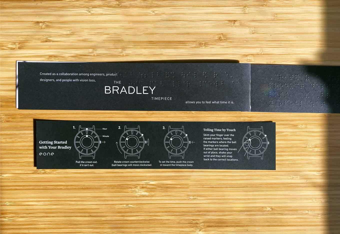 Boekje met de tekst "Gemaakt als een samenwerking tussen ingenieurs, productontwerpers en mensen met gezichtsverlies. Met het Bradley Timepiece kun je voelen hoe laat het is." Geschreven in braille.