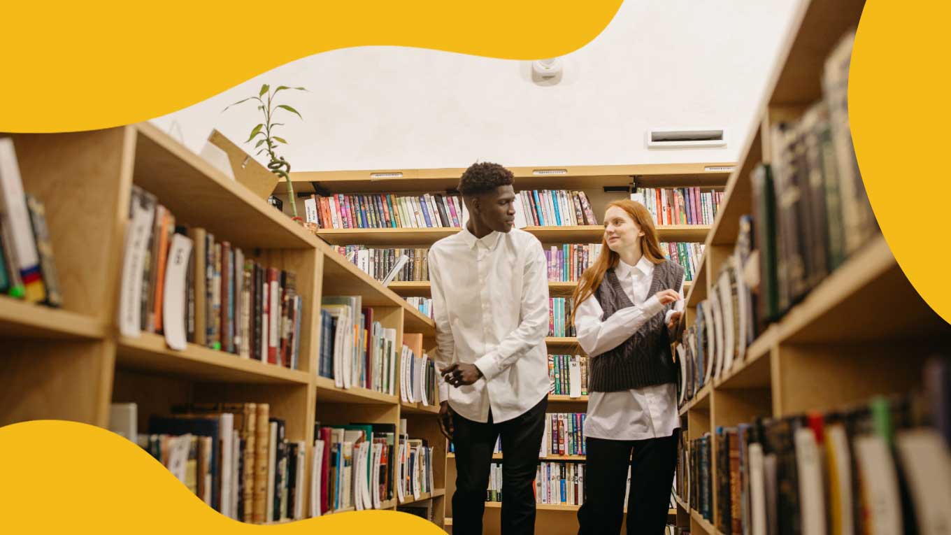 Een jonge zwarte man in een wit overhemd en zwarte broek praat met een jong wit meisje met rood haar en een grijs truivest. Ze lopen door de boekenkasten in een bibliotheek.