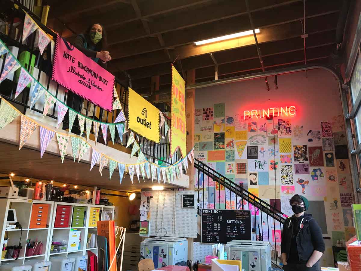 Een kleurrijke kamer vol prints en zines. Aan de muur hangt een roze neonbord "drukkerij." Twee mensen staan in de kamer met maskers op.