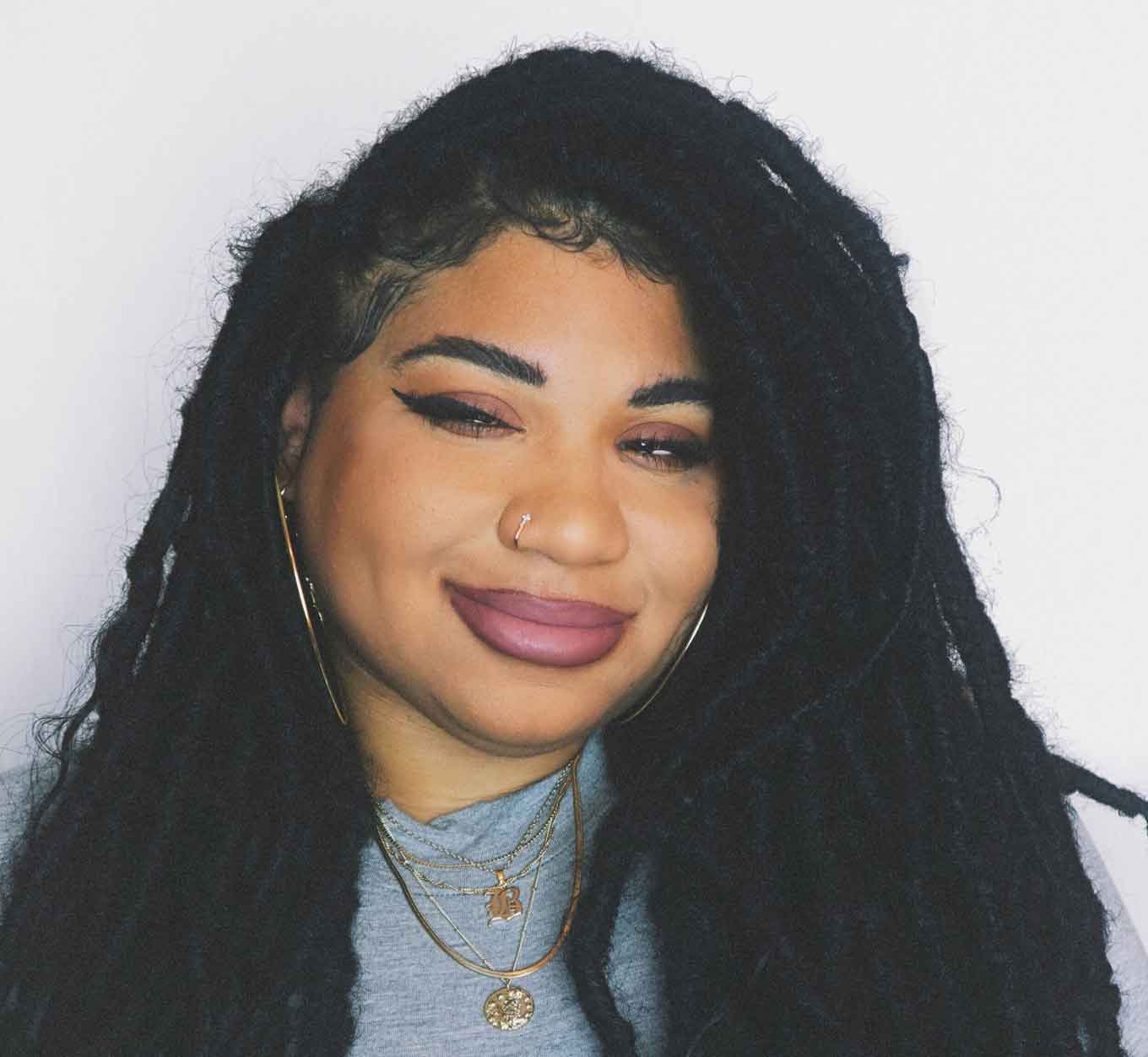 Een zwarte vrouw met dreadlocs draagt een grijs overhemd en gouden kettingen. Ze heeft roze lippenstift op en grijnst naar de camera. 