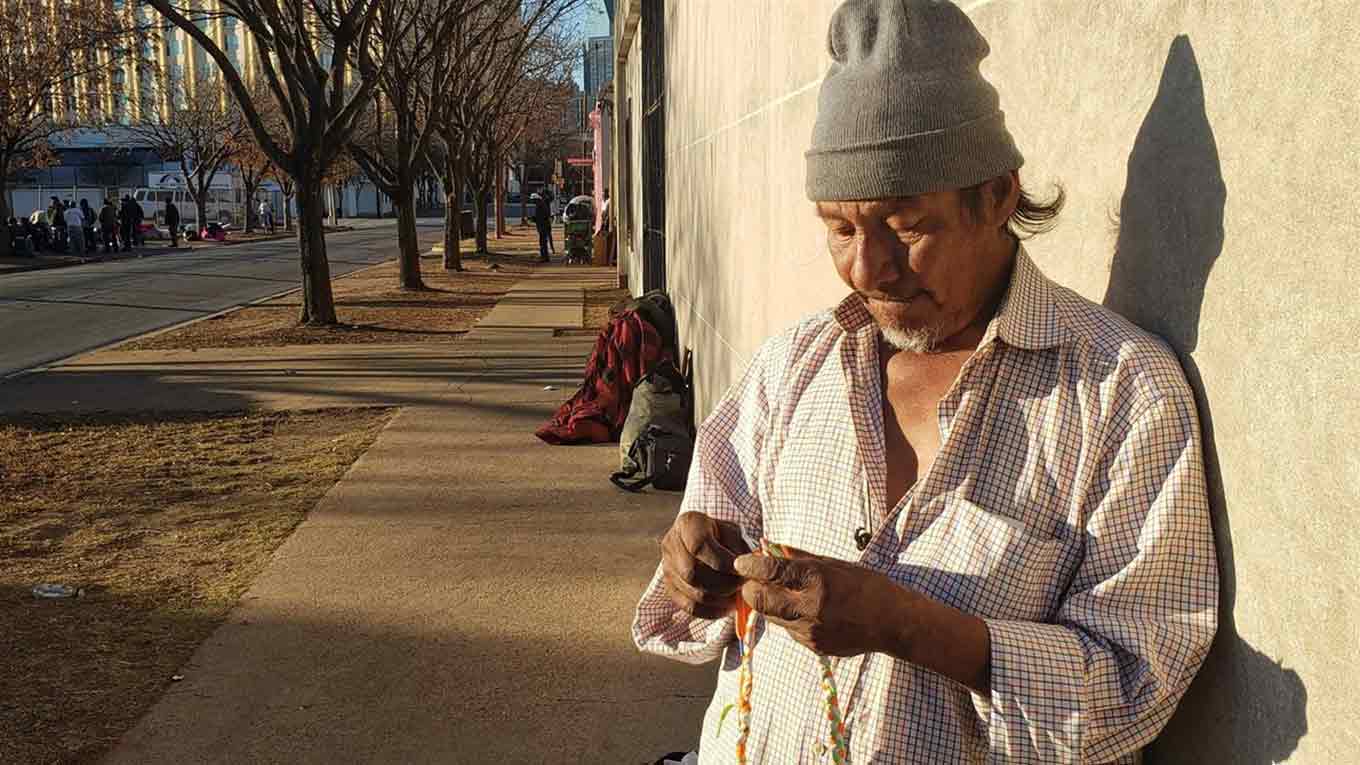 Een dakloze man kijkt neer op een vlecht die hij aan het doen is terwijl hij op een zonnige muur leunt.