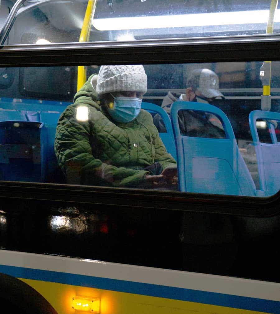 Een persoon zit in een bus, draagt een masker en een warme hoed, met behulp van zijn telefoon