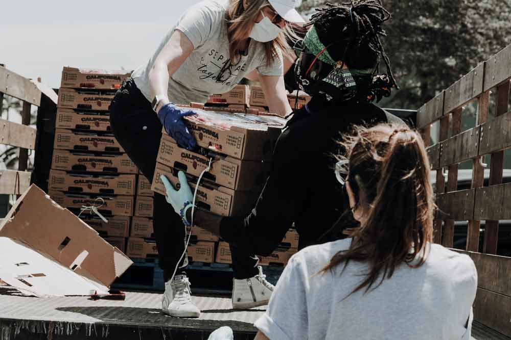 Een vrouw met een mondkapje staat achterop een vrachtwagen en overhandigt een vrijwilliger een doos met producten.
