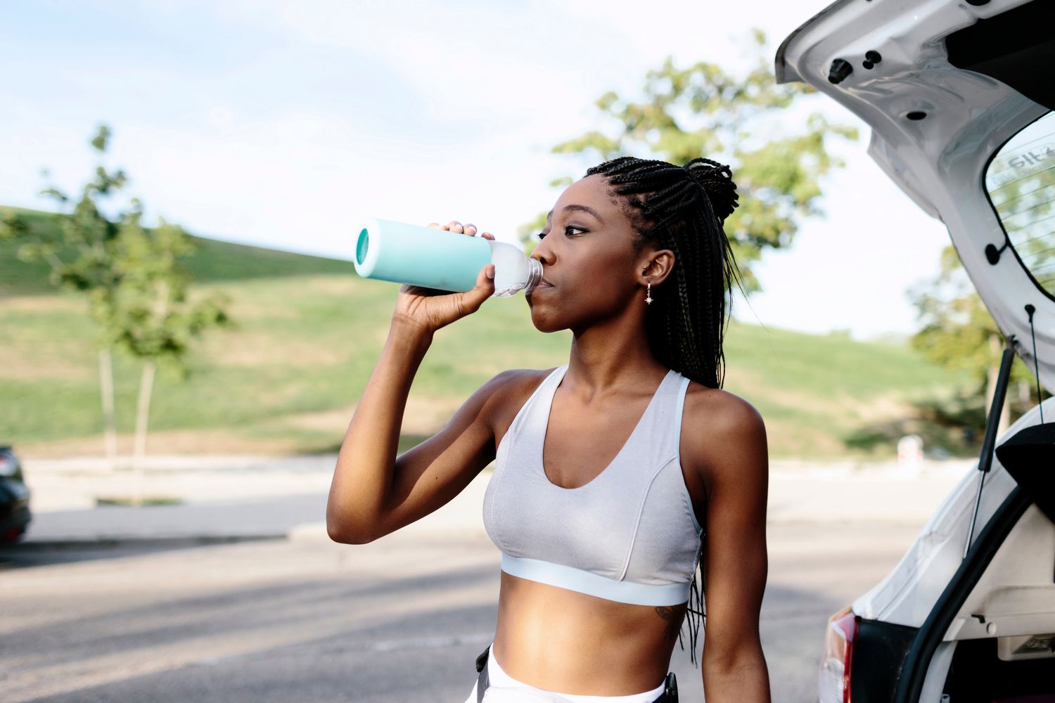 Aantrekkelijke jonge zwarte vrouw op sportkleding en met gevlochten kapsel drinkt water naast een auto voordat ze aan haar training begint. Overdag is ze overdag in een zomerochtend buiten naast een park.