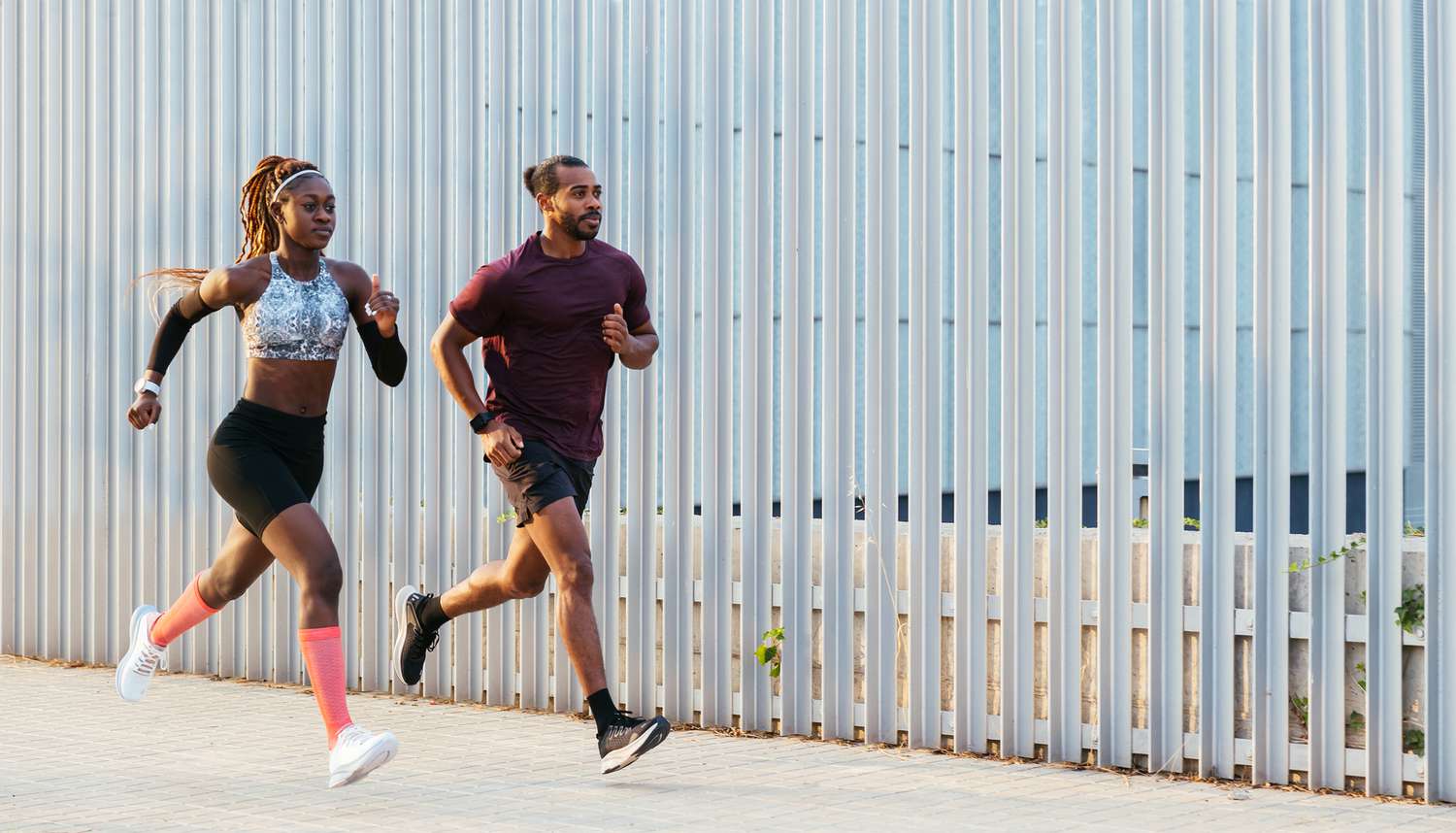 Volledig lichaam van zelfverzekerde gemotiveerde jonge Afro-Amerikaanse mannelijke en vrouwelijke hardlopers in sportkleding die samen trainen en snel rennen op de stoep bij het hek tijdens buitentraining